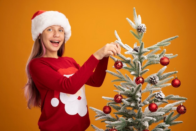 Bonne petite fille en pull de noël et bonnet de noel suspendu des boules sur un arbre de noël souriant joyeusement sur fond orange