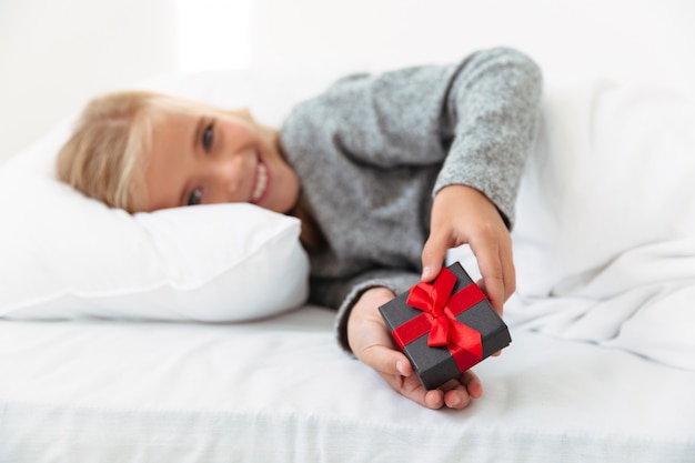 Bonne petite fille tenant une petite boîte cadeau en position couchée dans son lit, selective focus