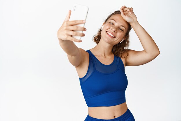 Bonne sportive prenant un selfie tout en essuyant la sueur, en faisant de l'exercice et en photographiant, debout en tenue de sport sur blanc.