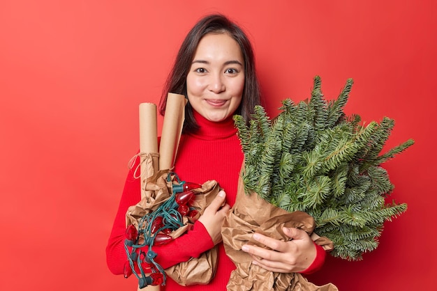 Photo gratuite bonnes fêtes de fin d'année. une jeune femme brune d'apparence orientale tient un bouquet de branches d'épinette en papier roulé et des guirlandes rétro préparent des décorations isolées sur fond rouge vif