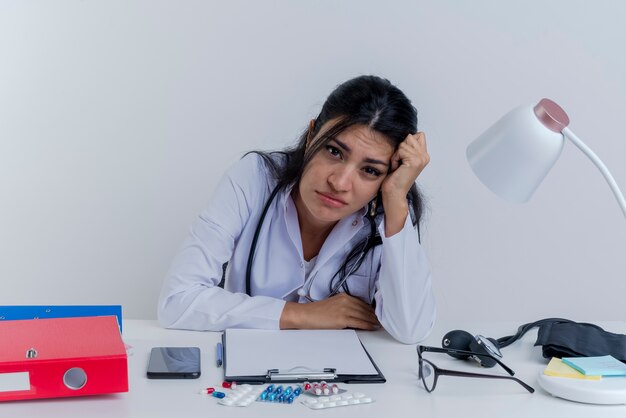 Bored jeune femme médecin portant une robe médicale et un stéthoscope assis au bureau avec des outils médicaux à la recherche de mettre les mains sur le bureau et sur la tête isolée