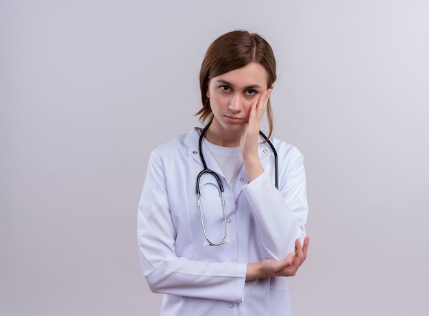 Bored jeune femme médecin portant une robe médicale et un stéthoscope et mettant la main sur la joue avec copie espace