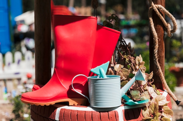 Botte en caoutchouc rouge; arrosoir et bêche près de la plante en pot