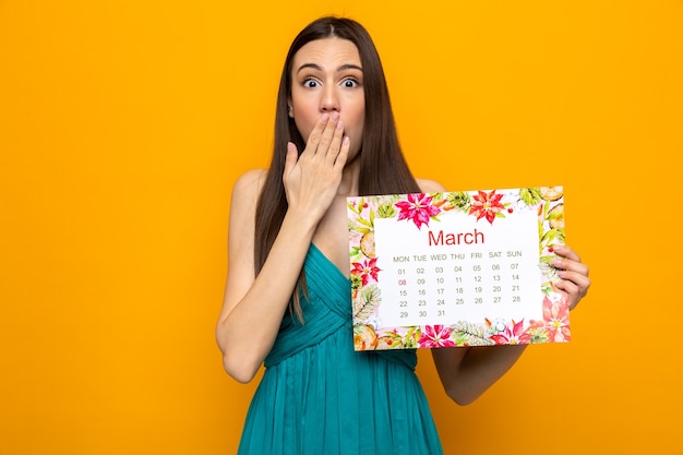 Bouche couverte de peur avec la main belle jeune fille le jour de la femme heureuse tenant le calendrier