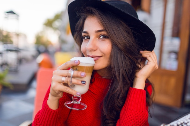 Bouchent la belle jeune femme brune au chapeau noir élégant et pull rouge vif assis dans un café en espace ouvert et boire du café avec du lait ou un cappuccino en matinée ensoleillée.