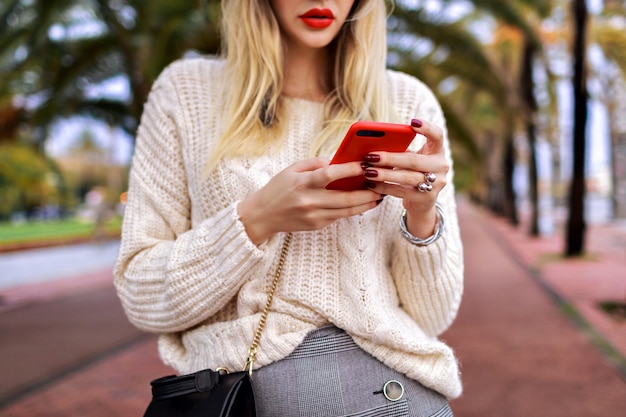 Bouchent les détails de la femme posant dans la rue et appuyez sur son smartphone, les lèvres rouges et le pull blanc à la mode confortable, la mode.