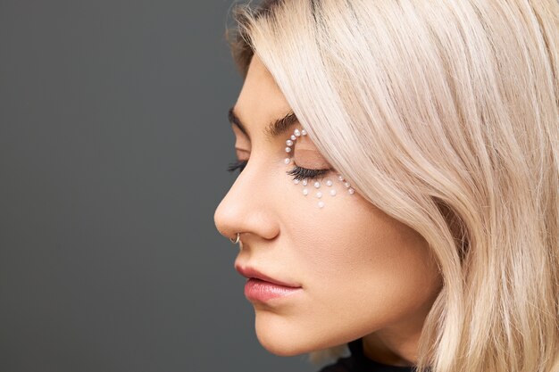 Bouchent l'image de profil de la belle jeune femme avec un maquillage parfait, une coiffure bob teinte, un anneau dans le nez et des cristaux blancs autour de ses yeux, posant isolé, gardant les yeux fermés. Beauté, soins de la peau et style