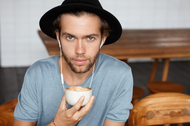 Bouchent le portrait de beau jeune homme aux yeux bleus avec une barbe élégante portant un chapeau noir et un t-shirt décontracté tenant une tasse de cappuccino et à la recherche tout en profitant de la bonne musique sur les écouteurs