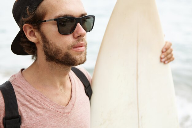 Bouchent le portrait de beau jeune homme barbu dans des lunettes de soleil élégantes posant à l'extérieur avec sa planche de surf blanche, profitant du magnifique paysage marin