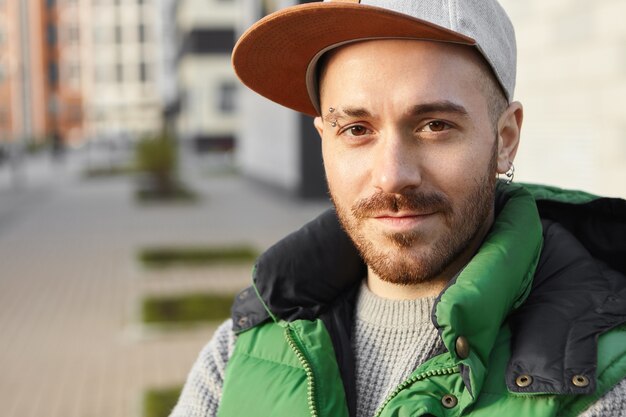 Bouchent le portrait de beau mec charismatique avec des yeux bruns, des soies et des boucles d'oreilles percées en attente de petite amie à l'extérieur avec le sourire. Concept de personnes, de mode de vie et de mode urbaine