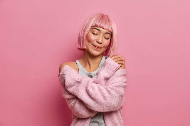 Bouchent le portrait de la charmante jolie femme asiatique a une coiffure rose, se serre dans ses bras, se tient les yeux fermés, porte un pull doux et chaud, se tient à l'intérieur