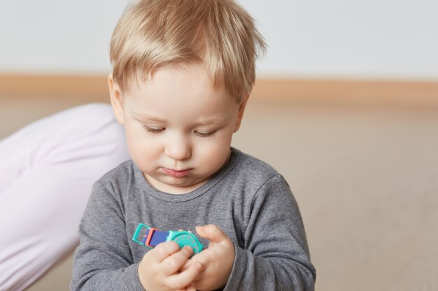 Bouchent le portrait d'enfant innocent regardant attentivement les montres-bracelets enfantines colorées à la maison. Petit garçon en chemise grise aux cheveux blonds. Le nourrisson aux joues potelées est devenu curieux du nouvel objet.