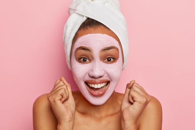 Bouchent le portrait d'une femme afro-américaine heureuse, serre les poings, applique un masque d'argile rose, obtient une thérapie de spa, porte une serviette enveloppée sur les cheveux mouillés