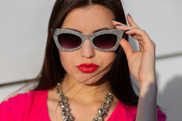 Bouchent le portrait d'une femme brune à la mode dans des lunettes de soleil élégantes et un t-shirt rose.