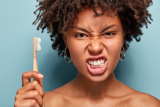 Photo gratuite bouchent le portrait d'une femme mécontente agacée fronce les sourcils, irrité par quelque chose, tient la brosse à dents, prend soin de l'hygiène bucco-dentaire, a une peau saine, isolée sur un mur bleu, nettoie les dents à l'intérieur