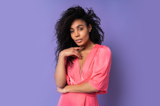 Bouchent le portrait d'une femme de race mixte confiante avec une coiffure frisée en robe rose debout sur un mur violet.