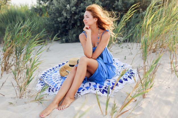 Bouchent le portrait d'une femme rousse souriante incroyable en robe bleue se détendre sur la plage ensoleillée de printemps sur une serviette. Chapeau de paille, bracelets et collier élégants.
