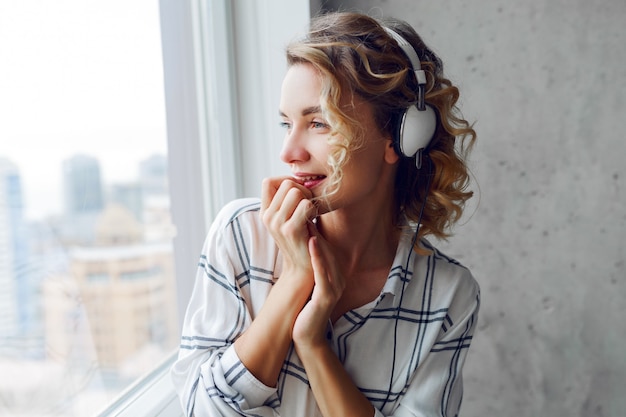 Bouchent le portrait d'une femme souriante pensive écoute de la musique par des écouteurs, posant près de la fenêtre. Intérieur urbain moderne.