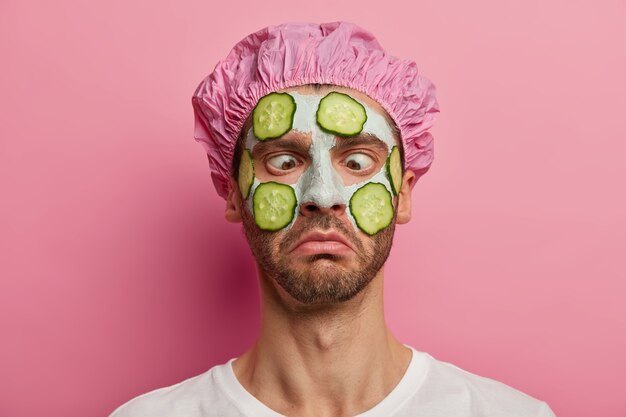 Bouchent le portrait d'un homme aux yeux croisés, désireux d'avoir une peau parfaite, applique un masque facial végétal et des concombres, porte un bonnet de douche