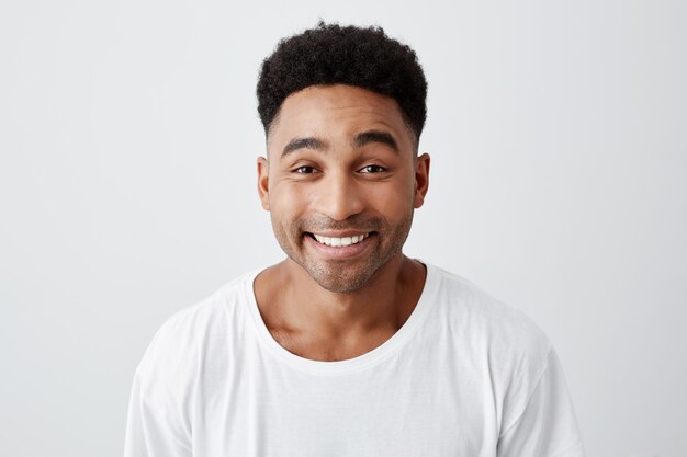 Bouchent portrait isolé de gai heureux jeune homme avec une coiffure afro en t-shirt blanc décontracté souriant brillamment, regardant à huis clos avec une expression excitée et joyeuse.
