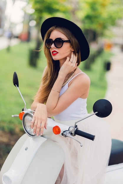 Bouchent le portrait de mode de jeune femme blonde en jupe de tulle blanc et talons noirs assis sur un scooter vintage.