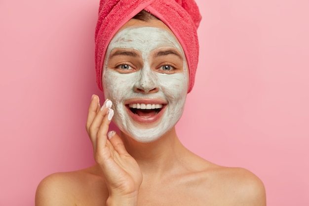 Bouchent le portrait d'un modèle féminin ravi heureux avec un masque facial cosmétique appliqué sur son visage, a des soins de beauté, porte une serviette enveloppée sur la tête, a une apparence saine et rafraîchie. Renouvellement et thérapie