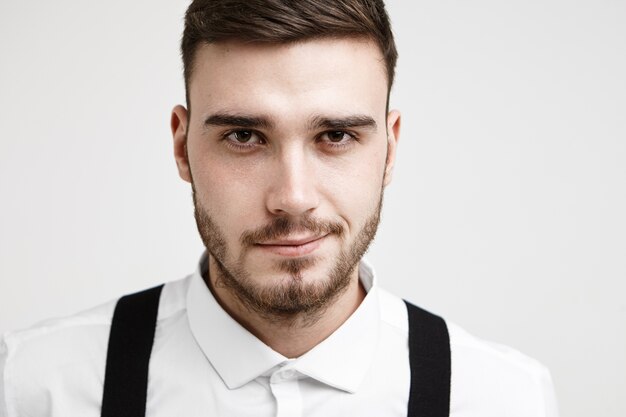 Bouchent le portrait de séduisant jeune homme brune avec une coiffure à la mode et une barbe taillée posant au studio blanc portant une chemise formelle et des bretelles, se sentant confiant quant à l'entretien d'embauche à venir