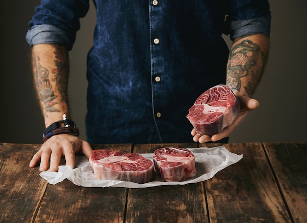 Un boucher brutal avec des mains tatouées offre un morceau de bon steak de viande crue à la caméra, d'autres steaks en papier kraft blanc sur une vieille table en bois vintage. méconnaissable