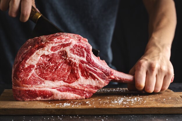 Boucher coupant un steak de tomahawk frais et appétissant sur une vieille planche de bois. Fermer