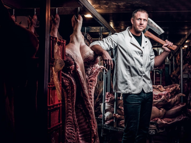 Le boucher dans une chemise de travail blanche tenant une hache debout dans un entrepôt réfrigéré au milieu de carcasses de viande