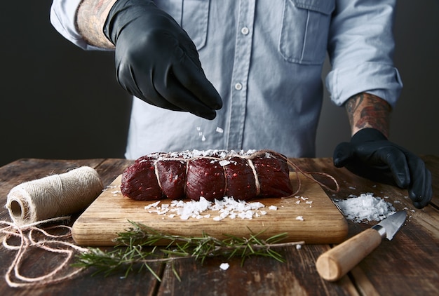 Photo gratuite boucher tatoué dans des gants noirs sels attaché un morceau de viande pour le fumer.