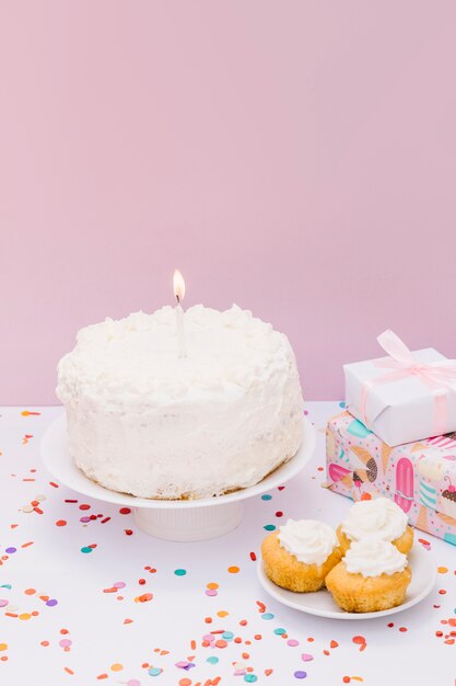 Bougie allumée sur un gâteau blanc avec des muffins et des cadeaux sur un bureau blanc contre un mur rose