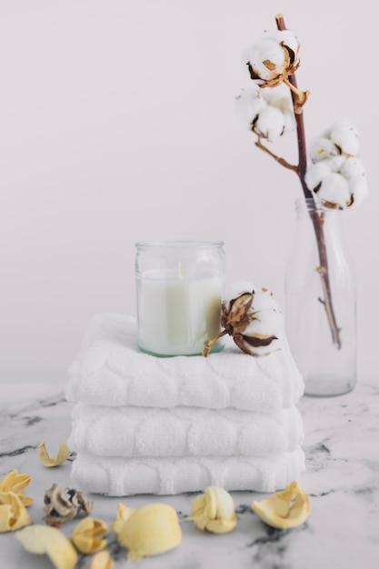 Bougie blanche dans un bougeoir sur des serviettes blanches empilées près des gousses sèches et une branche de coton en bouteille