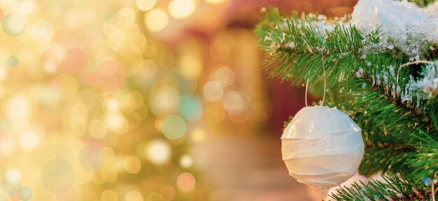 Boule de Noël blanche accrochée à une branche de sapin enneigé, fond effet bokeh