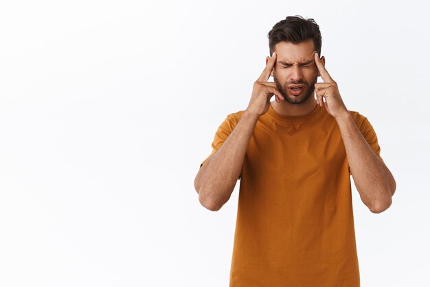 Bouleversé et affligé jeune barbu tendu souffrant de terribles migraines massant les tempes avec l'index fermer les yeux et grimaçant de douleur maux de tête douloureux debout fond blanc