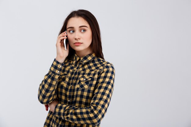 Bouleversé la belle jeune fille vêtue d'une chemise à carreaux parlant au téléphone, regardant de côté sur le mur blanc.