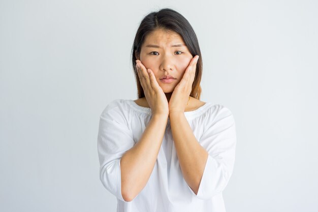 Bouleversé choqué jeune femme asiatique touchant les joues et regardant la caméra.