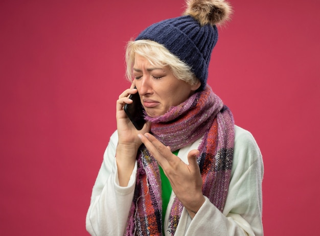 Bouleversé femme malsaine malade avec les cheveux courts dans une écharpe chaude et un chapeau se sentant mal à pleurer tout en parlant au téléphone mobile debout sur le mur rose