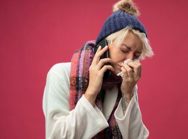 Photo gratuite bouleversé femme malsaine malade avec les cheveux courts dans une écharpe chaude et un chapeau se sentant mal pleurer tout en parlant au téléphone mobile en essuyant son nez avec une serviette debout sur fond rose