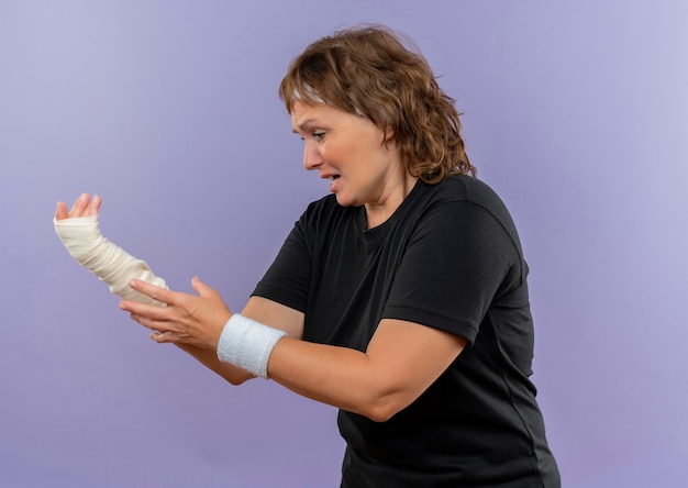 Photo gratuite bouleversé femme sportive d'âge moyen en t-shirt noir avec bandeau tenant son poignet bandé ayant des douleurs debout sur le mur bleu