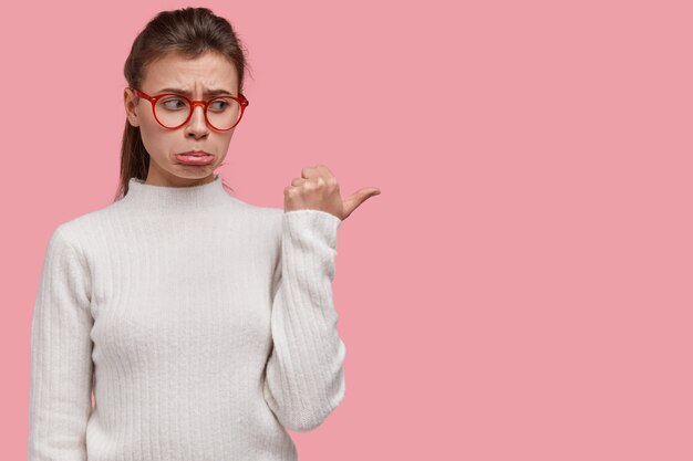 Bouleversé frustré jeune femme porte-monnaie lèvre inférieure, pointe avec le pouce à droite, montre quelque chose de négatif sur l'espace libre, porte un pull blanc