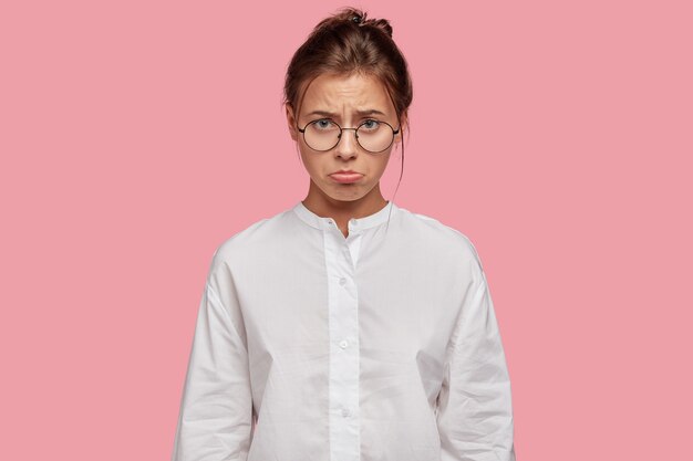 Bouleversé jeune femme avec des lunettes posant contre le mur rose