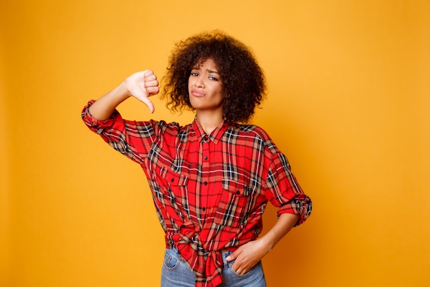 Bouleversé la jeune femme noire debout sur fond orange, les pouces vers le bas. Porter une chemise rouge et un jean.