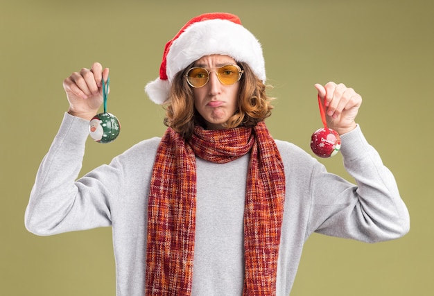 Bouleversé jeune homme portant bonnet de noel de Noël et des lunettes jaunes avec écharpe chaude autour de son cou tenant des boules de Noël regardant la caméra avec une expression triste pincer les lèvres debout sur fond vert