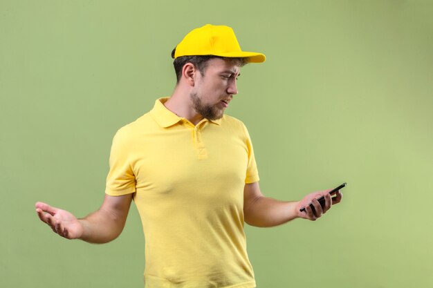 Bouleversé jeune livreur en polo jaune et casquette regardant l'écran de son smartphone répandre les mains à l'incertain et confus debout sur vert isolé