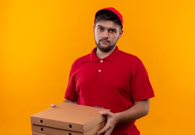 Bouleversé jeune livreur en uniforme rouge et cap holding pile de boîtes de pizza regardant la caméra avec une expression triste sur le visage