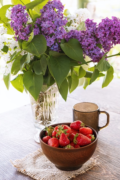 Bouquet de branches lilas dans un vase en cristal, bol en argile avec fraise rouge et tasse en verre foncé sur table en bois.