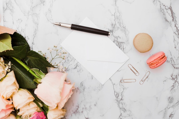 Bouquet de fleurs; stylo à plume; enveloppe; trombone et macarons sur fond texturé en marbre