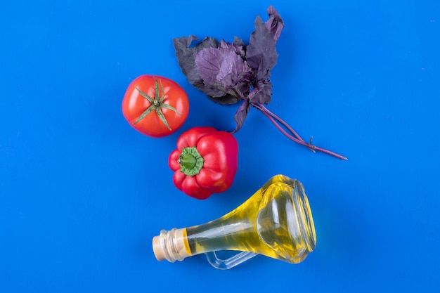 Bouteille en verre d'huile extra vierge avec basilic, poivron rouge et tomate sur table bleue.