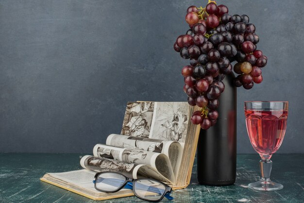 Bouteille de vin et grappe de raisin noir sur table en marbre avec livre et verres.